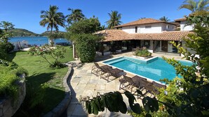 Un espace aménagé au bord de la piscine offrant une vue dégagée sur la mer, une maison avec un toit en tuiles et une pelouse bien entretenue.