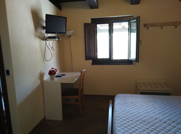 Una habitación pequeña con una cama, un escritorio con silla, un televisor montado en la pared y una ventana con persiana abierta.