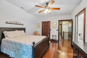A bedroom with a bed, a ceiling fan, a wooden floor, a door, and a mirror.