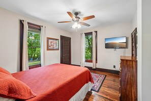 A bedroom with a bed, a ceiling fan, a television, and a view of greenery outside.