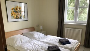 En pænt redt seng med hvide sengetæpper, et foldet håndklæde og et indrammet billede af solsikker på væggen.