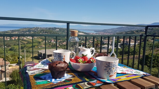 Un tavolo con una tovaglia colorata, una brocca, due tazze, una ciotola di fragole e un muffin al cioccolato su un balcone con vista su un paesaggio suggestivo.