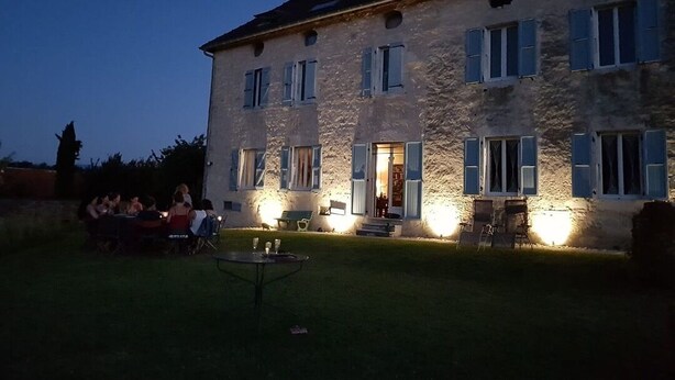 A stone building with multiple windows and a well-lit entrance. There is a group of people gathered outside, and a small table with chairs in the foreground.
