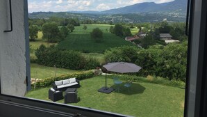 A view from a window overlooking a lush green landscape with a grassy lawn, a patio area with outdoor furniture, and a mountain range in the distance.
