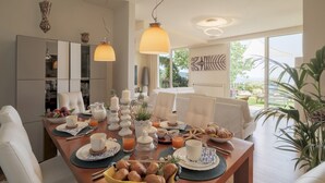 A modern dining area with a wooden table set for breakfast, featuring pastries, fruits, and beverages.