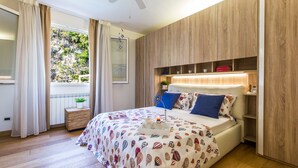 A bedroom with a bed, a bedside table, a nightstand, a window with curtains, and a view of greenery outside.