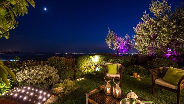 An outdoor seating area with a view of the city at night, including a wooden bench, chairs, and a table with a lantern.