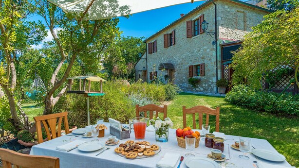 An outdoor dining area with a table set for a meal, surrounded by greenery and a stone building in the background. - La Casa dell'Artista 6, Emma Villas (Asciano)