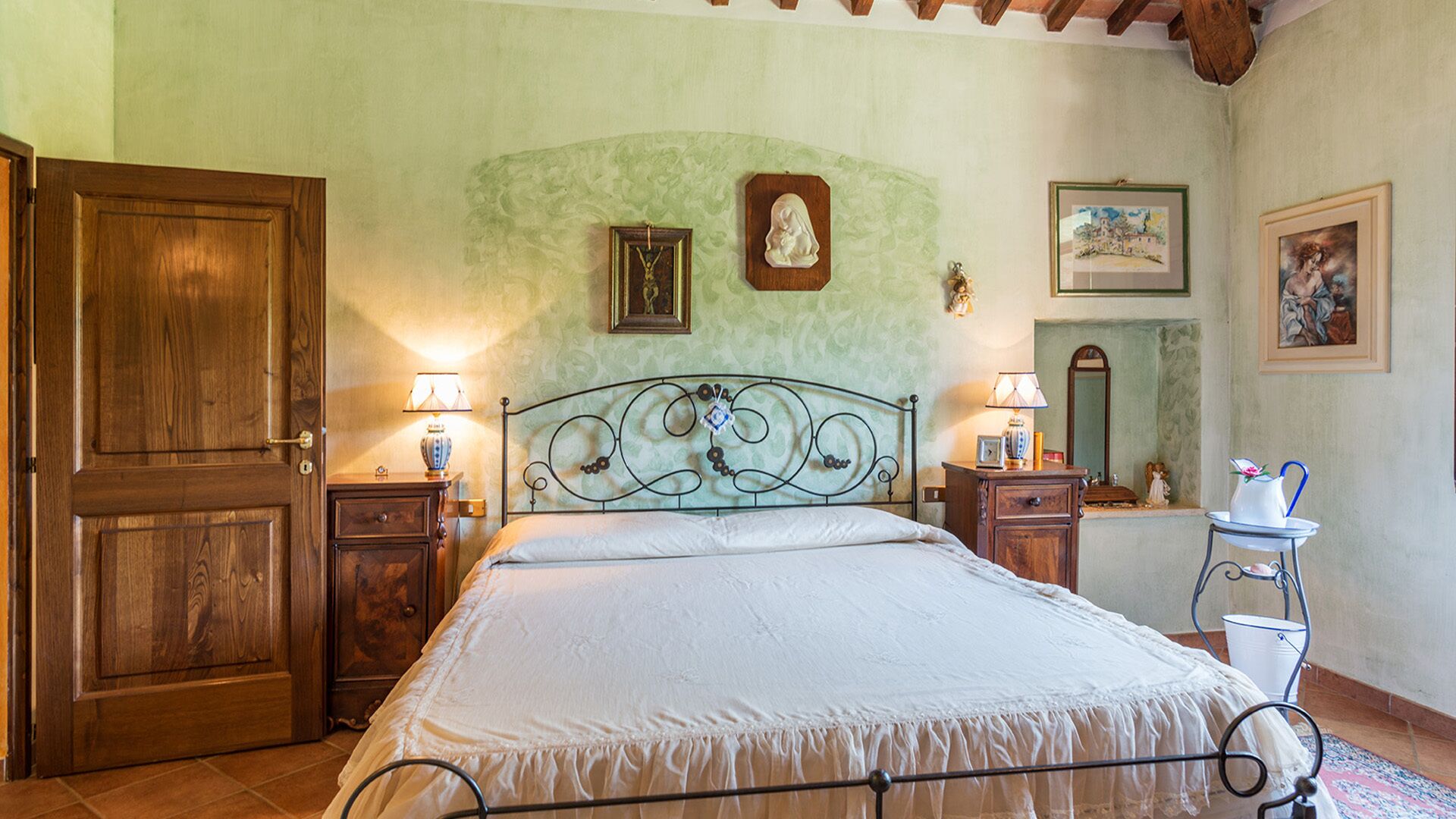 Une chambre avec une porte en bois, un lit avec un cadre en métal, deux tables de chevet avec des lampes et une petite table sur laquelle se trouve une carafe.