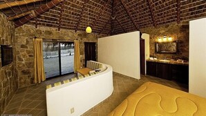 Une pièce aux murs en pierre, avec un plafond en bois, un canapé, une fenêtre avec des rideaux et une salle de bain équipée d’un lavabo et d’un miroir.