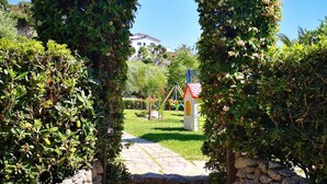 A stone pathway leading through a lush green archway to a playground. - Deluxe three-room apartments - at Le Casette sul Mare (Capo Vaticano)