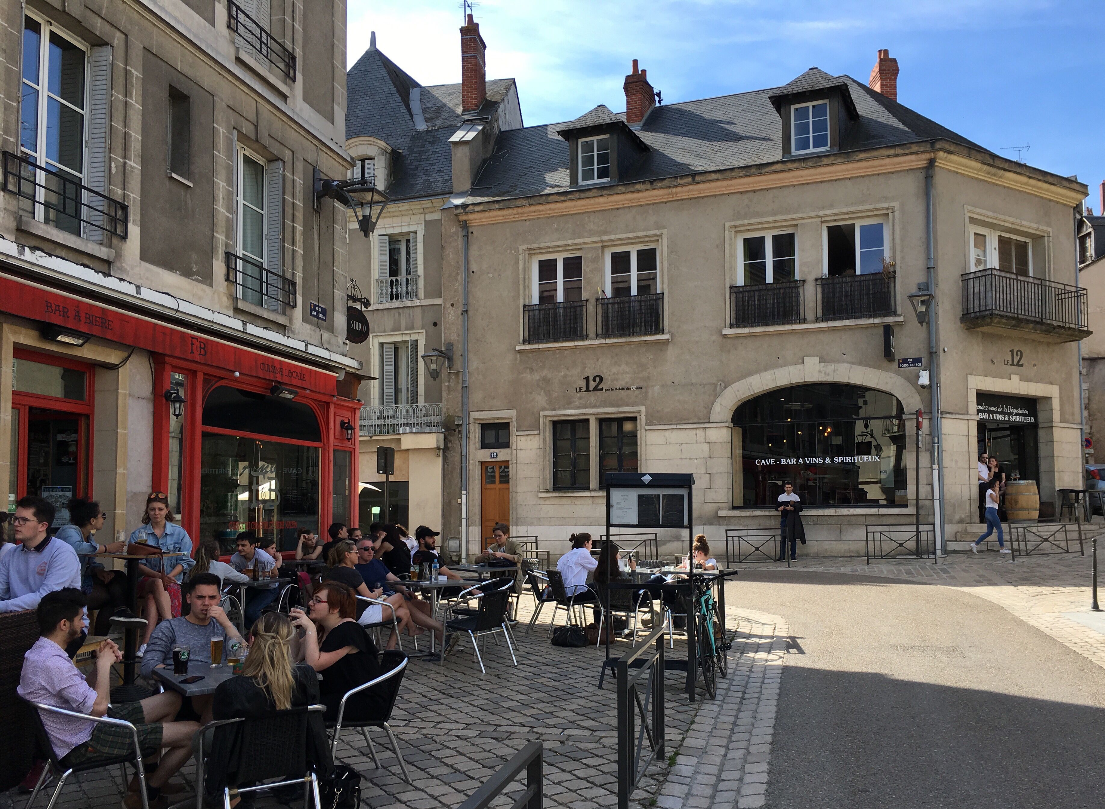 An outdoor café with tables and chairs, a bicycle, and a building with a sign 'CAVE - BAR A VINS & SPIRITEUX' in the background.