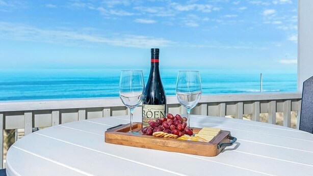 A table with a bottle of wine, two glasses, grapes, and cheese on a tray, overlooking a beach. - SC303 Seaside Dream by First Flight Rentals (Kill Devil Hills)