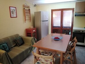 A compact living space with a sofa, a dining table, a refrigerator, a small kitchen area with a stove, and a window with blinds.
