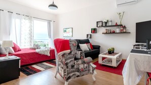 A modern living room with a red sofa, a patterned armchair, a black couch, and a television. There are shelves with decorative items and a window with curtains. - Apartment Anny1magnificant views over the ocean, mountains and banana plantation (Madeira)
