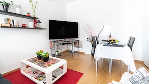 A modern living room with a flat-screen TV, a dining table with chairs, a coffee table with magazines, and a bookshelf with decorative items. - Apartment Anny1magnificant views over the ocean, mountains and banana plantation (Madeira)