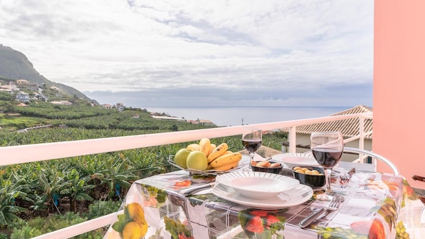 A table set with fruits, wine, and bread on a balcony overlooking a coastal landscape. - Apartment Anny1magnificant views over the ocean, mountains and banana plantation (Madeira)