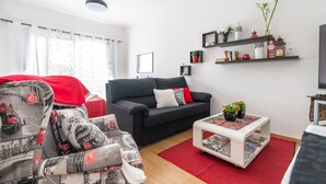A modern living room with a black sofa, a white coffee table with magazines, and a red rug. There are shelves with decorative items and a television on the wall. - Apartment Anny1magnificant views over the ocean, mountains and banana plantation (Madeira)