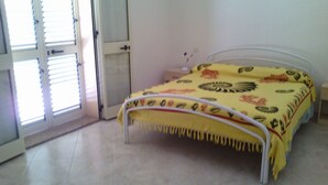 A single bed with a yellow bedspread in a room with a white door and a window with blinds. - House 2 rooms +Garden, barbecue, children max 11 years free, in Costa Verde.  (Fluminimaggiore)