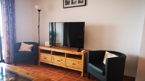 Una sala de estar con televisor, mueble de madera para entretenimiento, dos sillones, una lámpara y una vitrina de vidrio con conchas marinas.