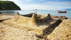 Uma praia arenosa com um castelo de areia grande e barcos ao fundo.