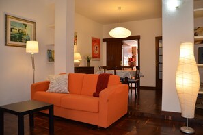 A living room with an orange sofa, a small table, and a tall floor lamp. There is a dining area in the background with a table and chairs.