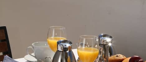 A breakfast setup on a balcony with a view of the sea, including a table with coffee cups, juice glasses, a sugar dispenser, pastries, and a fruit bowl.