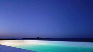 A rectangular infinity pool with clear water, surrounded by a white deck, under a twilight sky. - Beachfront villa with pool and spectacular views of Capo Passero Island (Portopalo di Capo Passero)