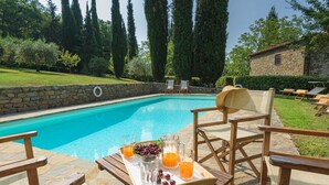 Une piscine bordée d’un mur en pierre, entourée de chaises longues en bois et d’une table sur laquelle se trouve un plateau de fruits et de boissons.