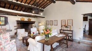 Un salon chaleureux avec des meubles blancs, une cheminée et un plafond en bois. On peut y voir des certificats encadrés accrochés au mur, ainsi qu’une bibliothèque remplie de livres.