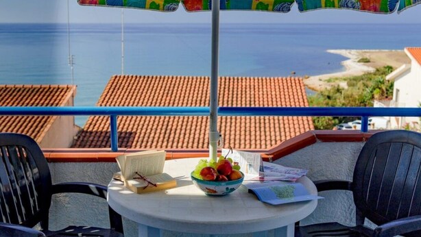 A balcony with a table, chairs, a bowl of fruit, a book, and an umbrella. - Apartment Lumia Standard (Sciacca)