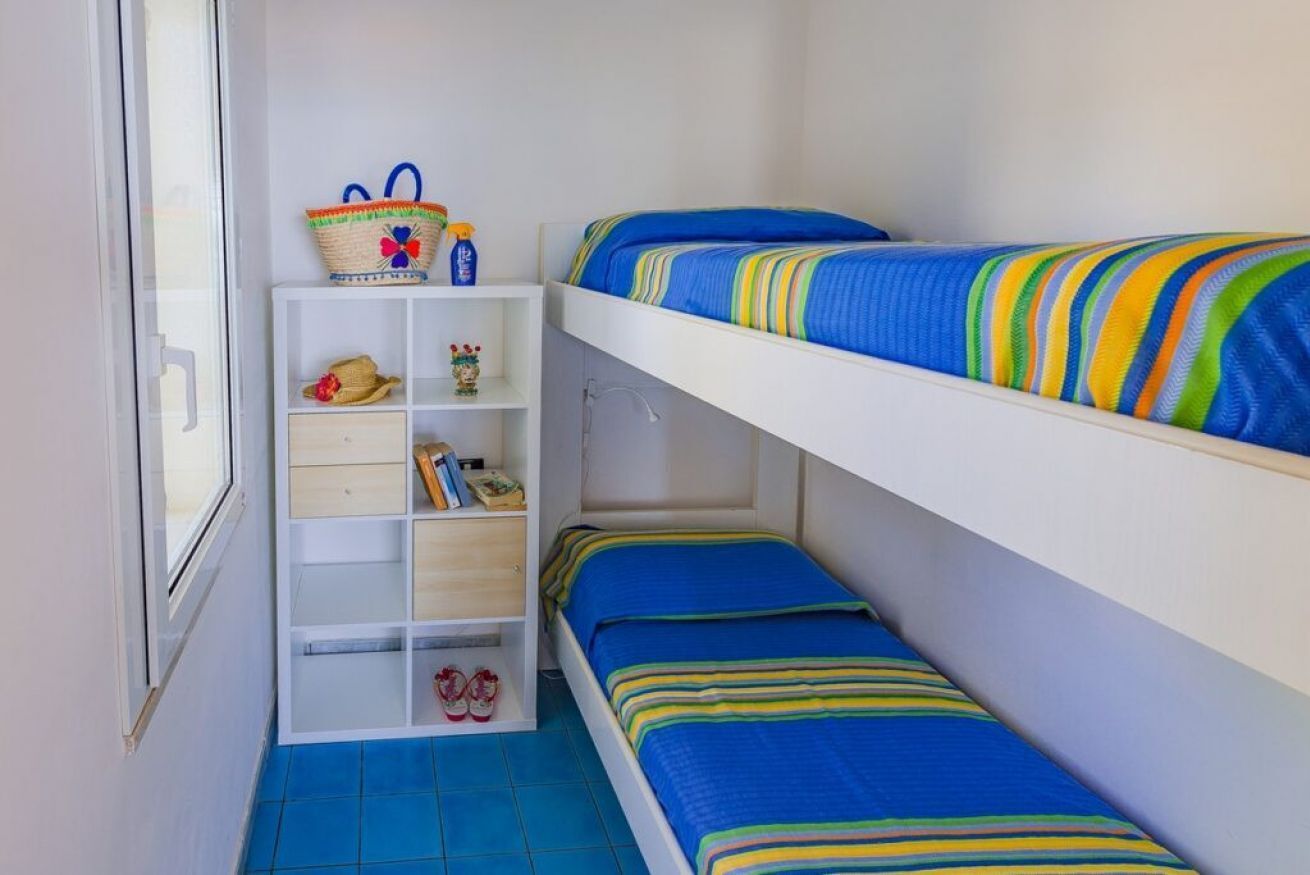 Una camera con letto a castello, un armadio bianco, uno scaffale con libri e un cesto, e una finestra.