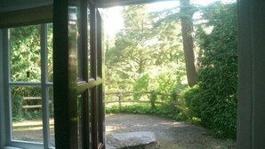 Vista desde una habitación a través de una ventana, mostrando un jardín con un muro de piedra, una valla de madera y abundante vegetación.