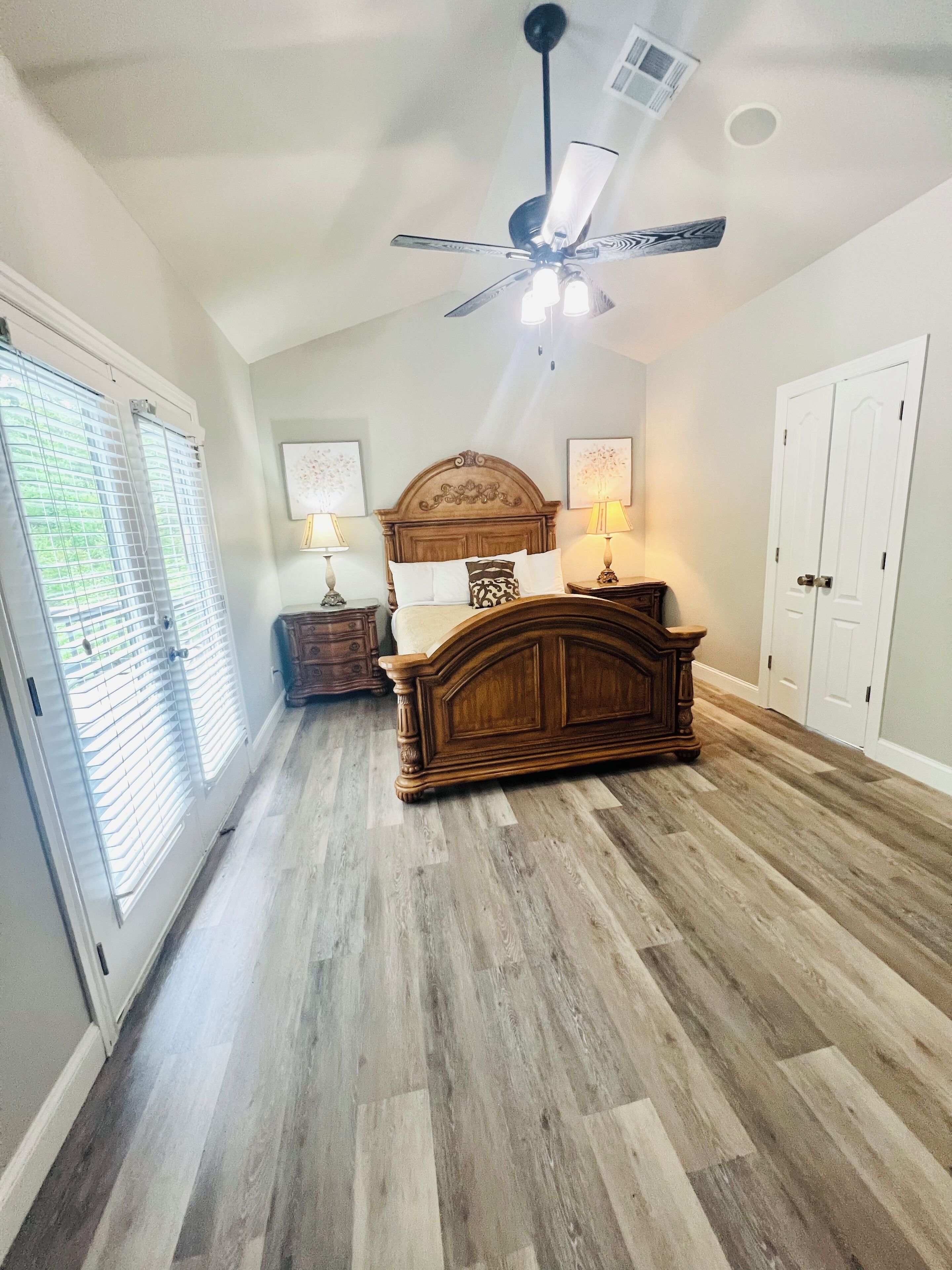 A bedroom with a wooden bed, two bedside tables with lamps, a ceiling fan, and a door with a window.