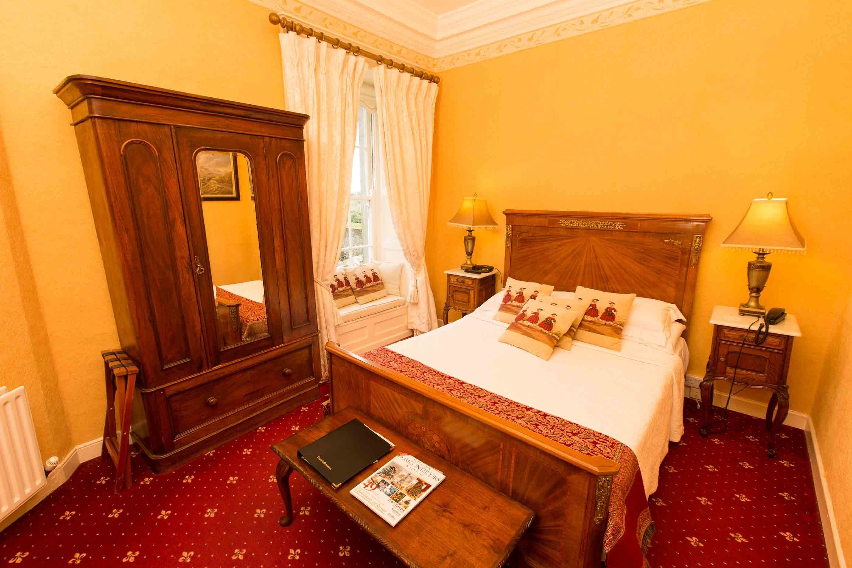 A hotel room with a wooden bed, a nightstand with a lamp, a dresser, and a window with curtains.
