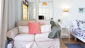 Una sala de estar moderna con un sofá, un escritorio pequeño y una cama. Hay dos espejos en la pared, una lámpara colgante y un estante con una planta.