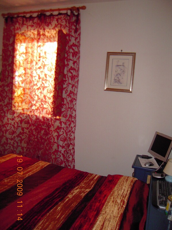 A bedroom with a bed, a desk with a computer, a framed picture on the wall, and a window with patterned curtains.