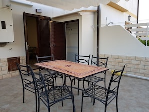 Um pátio com mesa e quatro cadeiras de azulejos, uma porta de madeira e uma parede branca com uma janela.