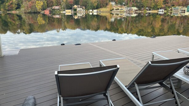 A wooden deck with lounge chairs and tables overlooking a calm lake, surrounded by autumn-colored trees and houses.