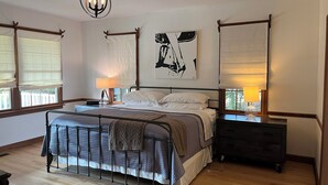A bedroom with a metal bed frame, a bedside table with a lamp, a nightstand, and a large window with blinds.