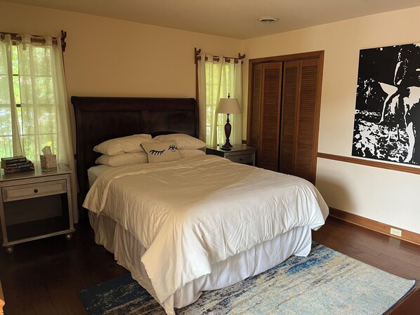 A neatly made bed with white linens, a bedside table with a lamp, a nightstand, and a window with sheer curtains.