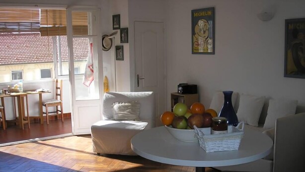 A living room with a white sofa, a round table with a bowl of fruit, a coffee table, and a television. There is a door with a coat hanging on it, and a window with blinds. - FESTIVAL SEA CLASS & CHARME CANNES MONTFLEURY (Cannes)