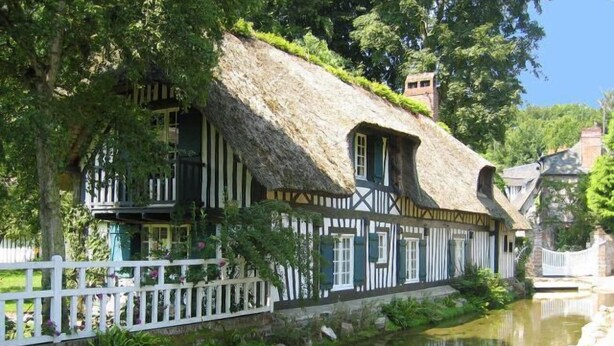 A traditional half-timbered house with a thatched roof, situated beside a calm river. - XVII century dovecote (Grémonville)
