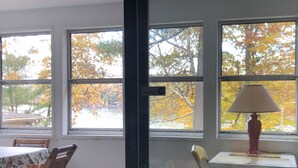 Una stanza con un tavolo da pranzo e sedie, una lampada su un tavolino e ampie finestre con vista sulle foglie autunnali.