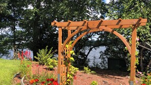 Un gazebo in legno con piante rampicanti, immerso in un giardino fiorito di fiori rossi e con vista su un lago.