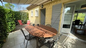 Un patio exterior con mesa de madera y sillas metálicas, una zona de estar techada y un cerramiento de vidrio corredero que da a un porche cubierto.