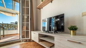 Ein modernes Wohnzimmer mit einem großen Flachbildfernseher, einer Holzkonsole und Blick auf einen Balkon mit Geländer sowie ein Gebäude im Hintergrund.