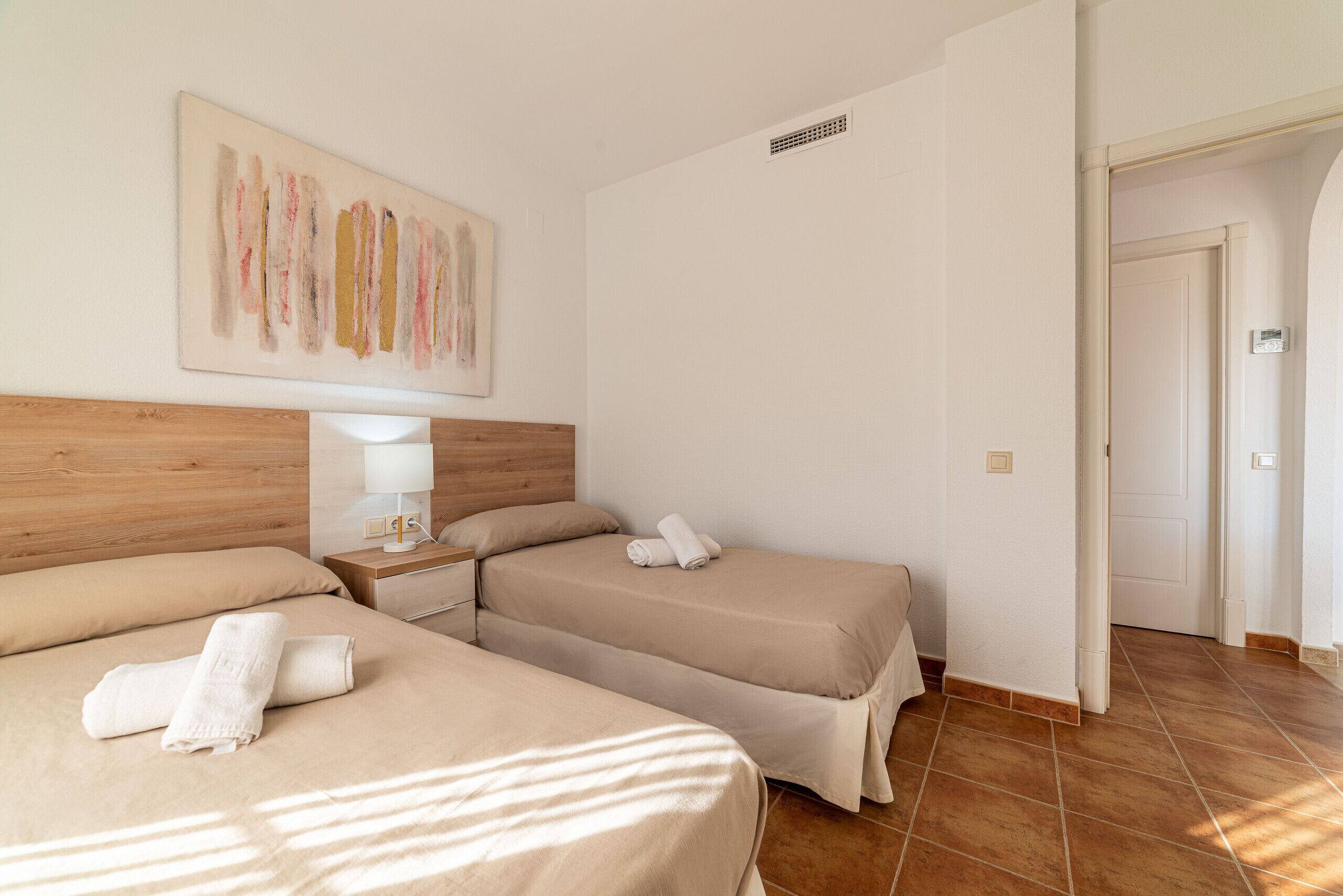 Habitación de hotel con dos camas, cabecera de madera, una obra de arte en la pared, una mesita de noche con lámpara y suelo de baldosas.