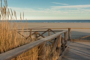 Une promenade en bois mène à une plage de sable bordée d’herbes hautes.