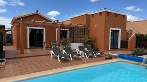 A swimming pool area with lounge chairs, a covered patio, and a building with a tiled roof. - Casa Piedra - Air Conditioned, Private Pool, Spa,  Pool Table, Frontline Golf (Caleta De Fuste)